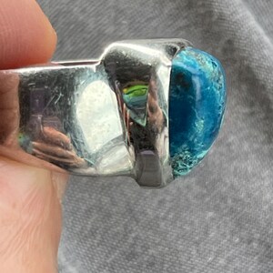 Puede incluir: Un anillo de plata con una gran piedra preciosa azul verdosa engastada en un bisel cuadrado. El anillo se lleva en un dedo.