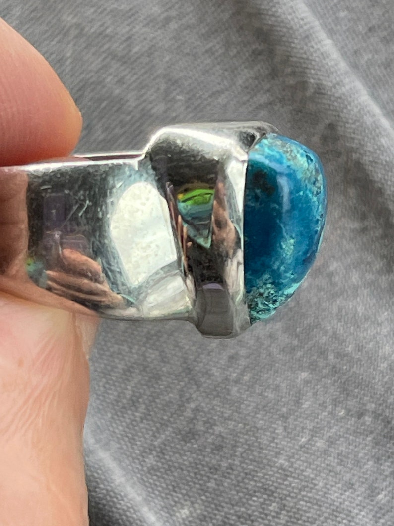 Puede incluir: Un anillo de plata con una gran piedra preciosa azul verdosa engastada en un bisel cuadrado. El anillo se lleva en un dedo.