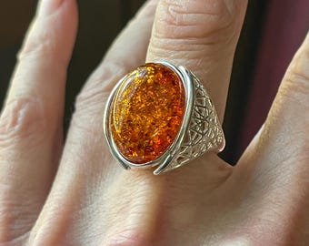 May include: A large, oval amber gemstone ring with a silver filigree band. The amber stone is a warm, golden-orange color and appears to have a textured surface. The ring is set in a detailed, openwork silver setting.