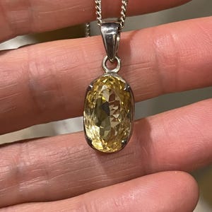 K&ouml;nnte beinhalten: Silberfarbene Halskette mit einem facettierten, oval geformten Anh&auml;nger aus gelbem Edelstein. Der Anh&auml;nger ist in einer silbernen L&uuml;nette gefasst und h&auml;ngt an einer filigranen Kette. Der Edelstein ist brillant geschliffen und reflektiert das Licht. Die Halskette ist ein Schmuckst&uuml;ck.