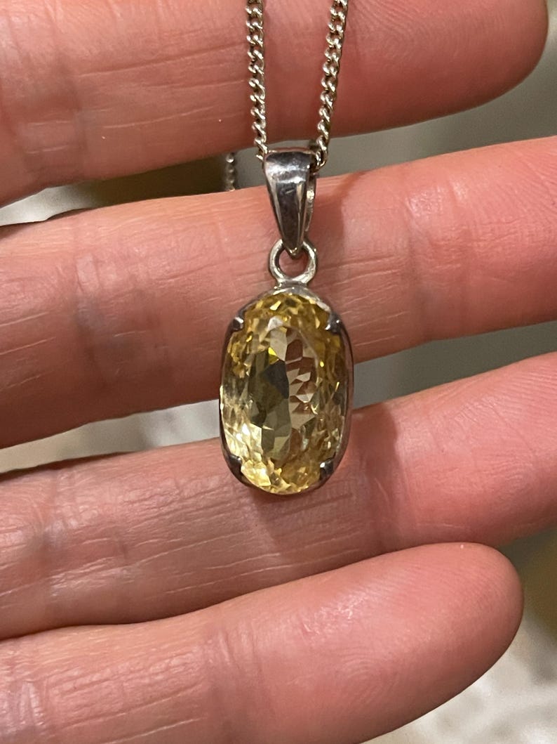 K&ouml;nnte beinhalten: Silberfarbene Halskette mit einem facettierten, oval geformten Anh&auml;nger aus gelbem Edelstein. Der Anh&auml;nger ist in einer silbernen L&uuml;nette gefasst und h&auml;ngt an einer filigranen Kette. Der Edelstein ist brillant geschliffen und reflektiert das Licht. Die Halskette ist ein Schmuckst&uuml;ck.