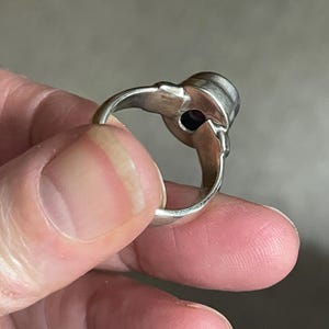 May include: A silver ring with a dark purple gemstone. The ring has a decorative design on the sides of the gemstone setting. The ring is held between fingers.