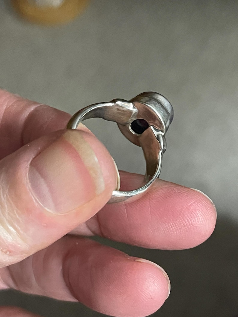 May include: A silver ring with a dark purple gemstone. The ring has a decorative design on the sides of the gemstone setting. The ring is held between fingers.