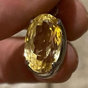 K&ouml;nnte beinhalten: Ein facettierter, oval geschliffener gelber Edelsteinanh&auml;nger, gefasst in einem silberfarbenen Metall. Der Edelstein hat einen brillanten, goldenen Farbton, und die Fassung scheint ein einfaches L&uuml;nettendesign zu sein. Der Anh&auml;nger wird zwischen den Fingern gehalten.