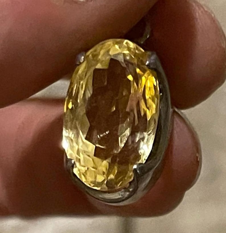 K&ouml;nnte beinhalten: Ein facettierter, oval geschliffener gelber Edelsteinanh&auml;nger, gefasst in einem silberfarbenen Metall. Der Edelstein hat einen brillanten, goldenen Farbton, und die Fassung scheint ein einfaches L&uuml;nettendesign zu sein. Der Anh&auml;nger wird zwischen den Fingern gehalten.