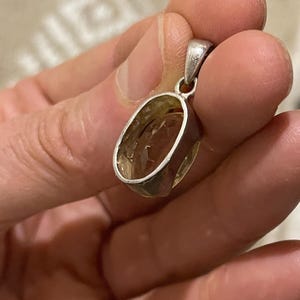 K&ouml;nnte beinhalten: Ovaler Anh&auml;nger mit einer silberfarbenen Metalleinfassung. Der Anh&auml;nger hat einen durchscheinenden, bernsteinfarbenen Stein. Die Fassung hat oben eine Schlaufe f&uuml;r eine Kette. Der Anh&auml;nger wird in einer Hand gehalten.