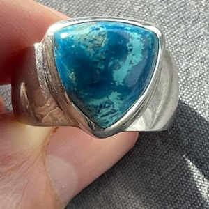 Puede incluir: Un anillo de plata con una piedra preciosa azul triangular engastada en el centro. El anillo tiene una banda texturizada.
