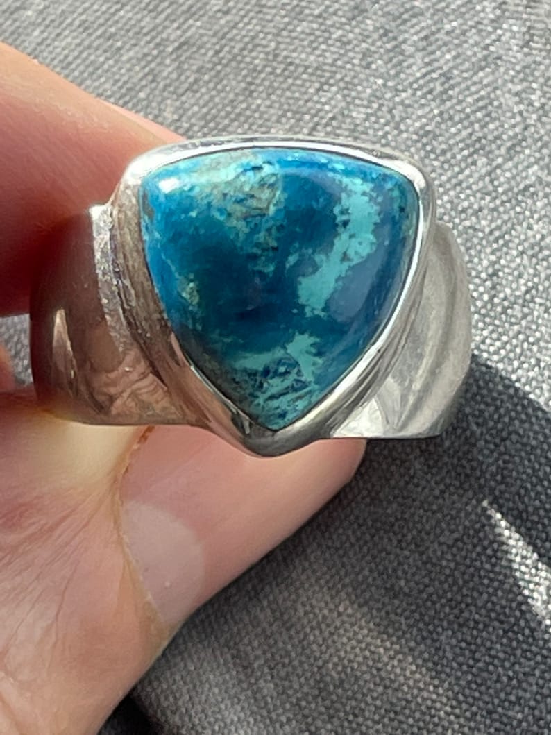 Puede incluir: Un anillo de plata con una piedra preciosa azul triangular engastada en el centro. El anillo tiene una banda texturizada.