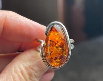 May include: A silver ring featuring a large, teardrop-shaped amber stone. The amber is a warm orange color with visible inclusions, set in a simple, elegant silver band. The ring is a statement piece.