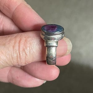 May include: A silver ring with a large, oval-shaped stone. The stone has a mix of colors, including blue and purple. The ring's band is a simple, polished silver. The ring is held up by a person's fingers.