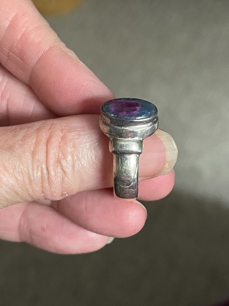 May include: A silver ring with a large, oval-shaped stone. The stone has a mix of colors, including blue and purple. The ring's band is a simple, polished silver. The ring is held up by a person's fingers.