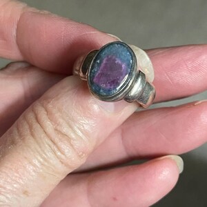 May include: A silver ring with an oval-shaped stone. The stone has a dark blue and purple color. The ring has a simple, elegant design. The ring is held by a person's hand.