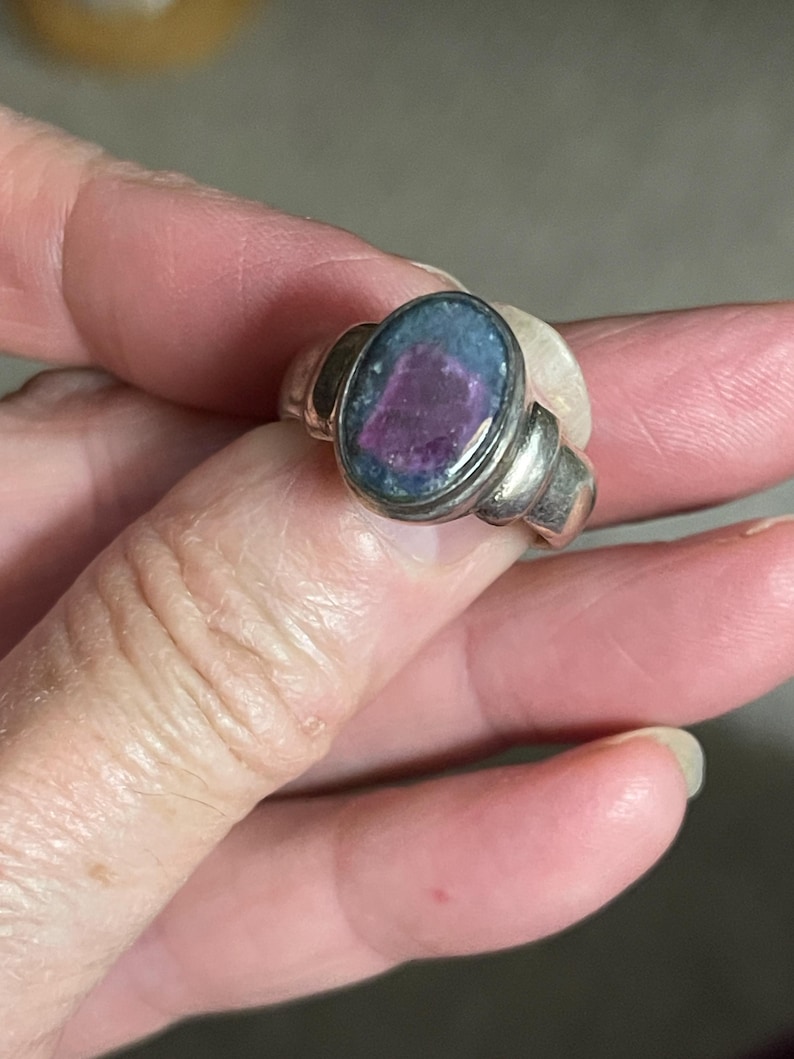 May include: A silver ring with an oval-shaped stone. The stone has a dark blue and purple color. The ring has a simple, elegant design. The ring is held by a person's hand.