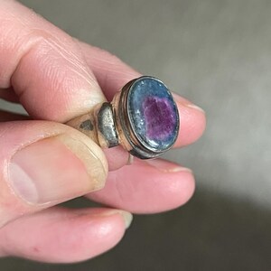 May include: Silver ring with an oval-shaped gemstone. The gemstone has a vibrant purple center surrounded by a blue-green border. The ring's band is a simple, classic design.
