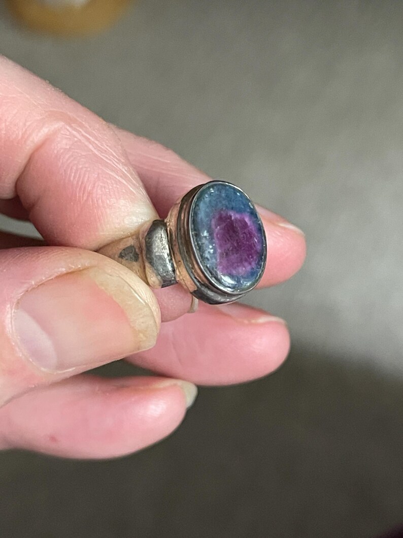 May include: Silver ring with an oval-shaped gemstone. The gemstone has a vibrant purple center surrounded by a blue-green border. The ring's band is a simple, classic design.