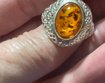 May include: A silver ring with an oval amber stone. The amber stone is a warm, golden-brown color with darker inclusions. The ring's band has a detailed, textured design around the stone, adding to its vintage appeal. The ring is a statement piece.