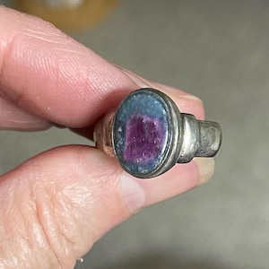 May include: A silver ring with an oval-shaped stone. The stone has a dark blue border and a central section of deep magenta color. The ring's band is a simple, classic design.