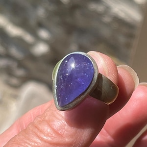 May include: A silver ring featuring a large, pear-shaped, deep purple gemstone. The gemstone is set in a silver bezel and has a polished, reflective surface. The ring's band is a simple, rounded design.