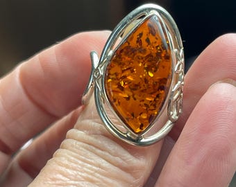 May include: A silver ring featuring a large, oval-shaped amber stone. The amber is a warm, translucent orange color with flecks throughout. The ring has a decorative silver setting with scrollwork details.
