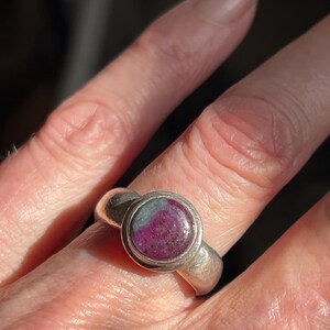 May include: A silver ring featuring a round, cabochon-cut gemstone with a mottled appearance, displaying deep magenta and dark green color variations. The stone is set in a smooth, polished silver bezel on a wide, plain silver band.