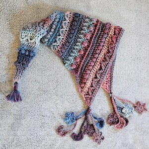 Könnte beinhalten: Ein handgefertigter Häkelschal in spitzer Form mit einem Farbverlauf von Blau über Lila bis Rosa. Der Schal hat eine Quaste am Ende und dekorative florale Akzente. Der Schal ist mit verschiedenen strukturierten Stichen gefertigt.