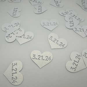 May include: A scattering of silver heart-shaped confetti with the date 3.21.24 printed on each piece.