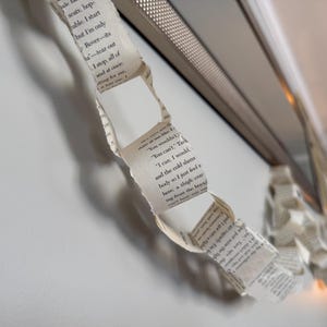 May include: A paper chain made from book pages hangs near a silver-colored frame. The chain is made of torn strips of paper with printed text. The background is a white wall.