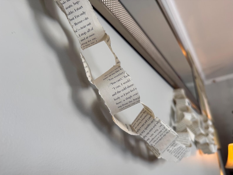 May include: A paper chain made from book pages hangs near a silver-colored frame. The chain is made of torn strips of paper with printed text. The background is a white wall.