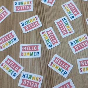 May include: Rectangular white stickers scattered on a wooden surface. Each sticker has the words "HELLO SUMMER" in colorful letters, with a red banner design. The stickers are a summer-themed product.