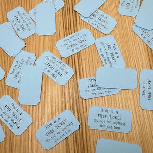 May include: A collection of light blue tickets scattered on a wooden surface. Each ticket has rounded corners and the text "This is a FREE TICKET. It's not for anything. It's just free." printed in black.