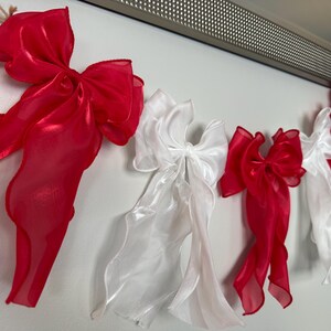 May include: A garland of red and white organza bows hangs against a white wall. The bows are tied with long, flowing ribbons. The garland is suspended from a natural twine.
