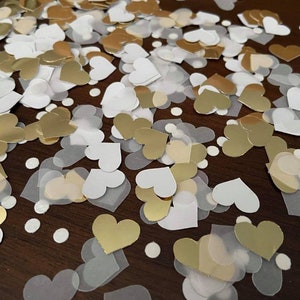 May include: A scattering of gold and white heart-shaped confetti on a brown wooden surface. The confetti is made of paper and foil.