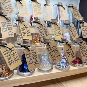 May include: A collection of small glass bottles with cork stoppers arranged on a tiered wooden stand. Each bottle contains various decorative items and features a brown paper tag tied with black string, labeled with names like Lunar Elixir and Snake Skin.