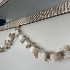 May include: A decorative garland made from folded book pages hangs below a silver framed mirror. The garland is made of connected paper loops, displaying printed text. The wall is painted a light gray color.