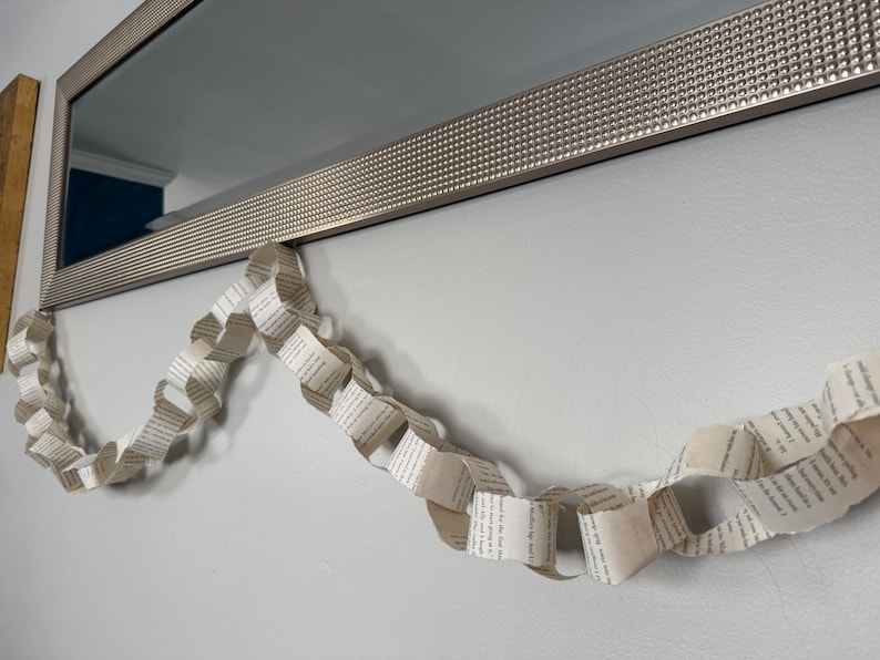 May include: A decorative garland made from folded book pages hangs below a silver framed mirror. The garland is made of connected paper loops, displaying printed text. The wall is painted a light gray color.