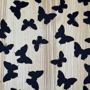 May include: A collection of flat, black butterfly-shaped cutouts scattered across a light-colored wood grain surface. Each butterfly silhouette features distinct wing shapes and antennae, arranged in a random pattern across the light background.