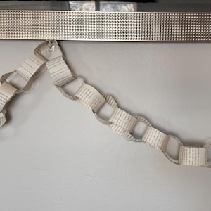 May include: A paper chain garland made from book pages, hanging against a light gray wall. The garland is made of interlocking loops of paper, with text visible on each page. A decorative silver trim is at the top.