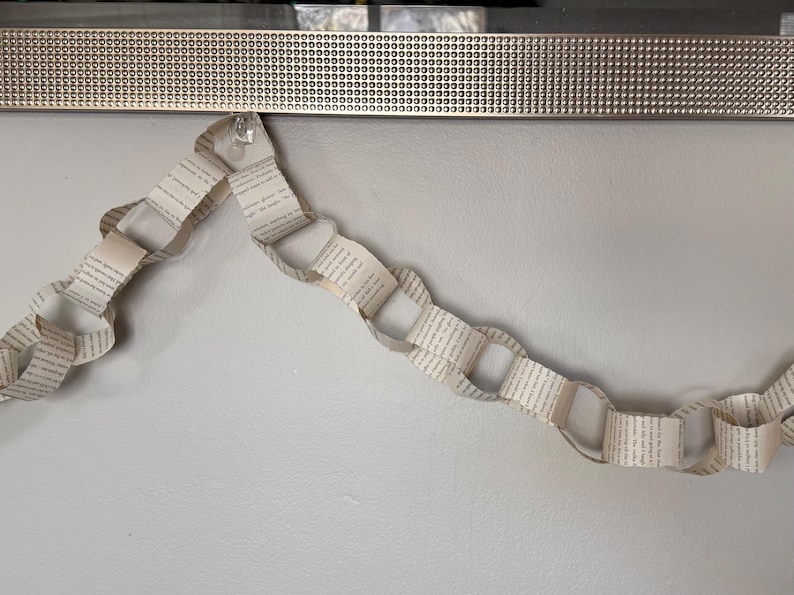 May include: A paper chain garland made from book pages, hanging against a light gray wall. The garland is made of interlocking loops of paper, with text visible on each page. A decorative silver trim is at the top.