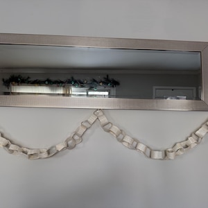May include: A rectangular mirror with a silver frame hangs on a gray wall. A decorative paper chain garland is suspended below the mirror. The mirror reflects a room with a decorated window.