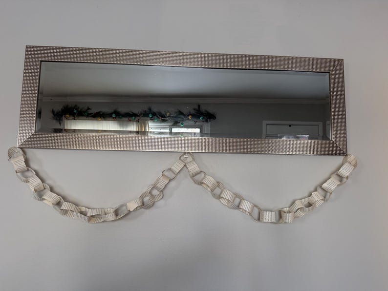 May include: A rectangular mirror with a silver frame hangs on a gray wall. A decorative paper chain garland is suspended below the mirror. The mirror reflects a room with a decorated window.
