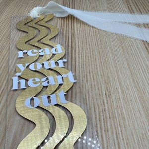 May include: A clear bookmark with a gold wavy design and the words "read your heart out" in white. It has a white ribbon attached at the top. The bookmark is on a wooden surface.