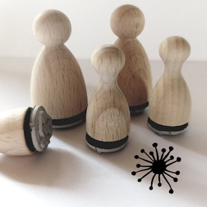 Könnte beinhalten: Vier Holzstempel mit schwarzen Stempelflächen. Die Stempel sind menschenförmig und in einer Reihe angeordnet. Der Stempel links liegt auf der Seite. Der Stempel in der Mitte ist der größte und der Stempel rechts ist der kleinste. Die Stempel sind alle aus hellem Holz und haben eine natürliche Oberfläche. Ein schwarzer Stempel mit einem stilisierten Sonnenstrahl befindet sich im Vordergrund.