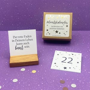 May include: A brown box with a white label that says "Adventskalender" and "24 K&auml;rtchen mit positiven Gedanken". There is a small wooden stand with a white card that says "Der rote Faden in Deinem Leben kann auch bunt sein." and a white card with black stars and the number 22.
