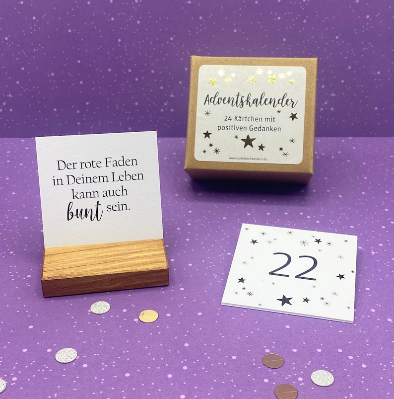 May include: A brown box with a white label that says "Adventskalender" and "24 K&auml;rtchen mit positiven Gedanken". There is a small wooden stand with a white card that says "Der rote Faden in Deinem Leben kann auch bunt sein." and a white card with black stars and the number 22.