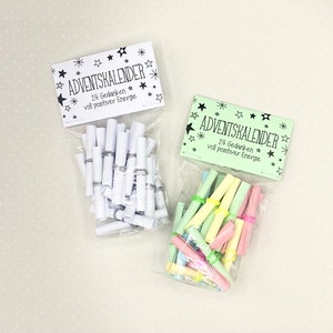 May include: Two clear plastic bags filled with rolled up pieces of paper. The bags have green and white labels that say "Adventkalender" and "24 Gedanken voll positiver Energie".