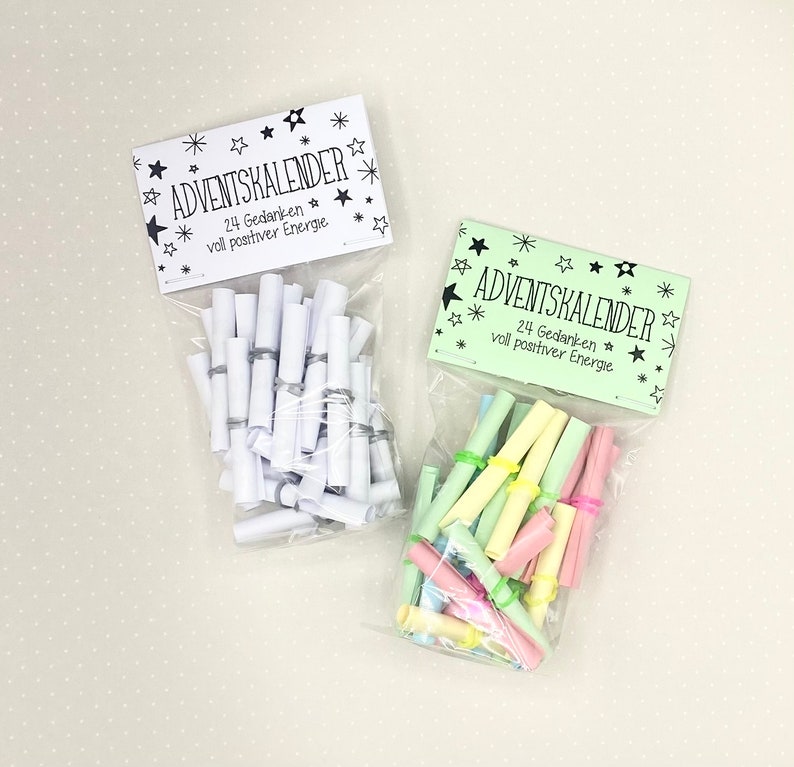 May include: Two clear plastic bags filled with rolled up pieces of paper. The bags have green and white labels that say "Adventkalender" and "24 Gedanken voll positiver Energie".