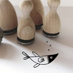 Könnte beinhalten: Vier Holzstempel mit schwarzen Stempelflächen, die die Form eines abgerundeten Kegels haben, sind auf einer weißen Oberfläche angeordnet. Unter den Stempeln ist eine Schwarzweißillustration eines Fisches mit Blasen gezeichnet.