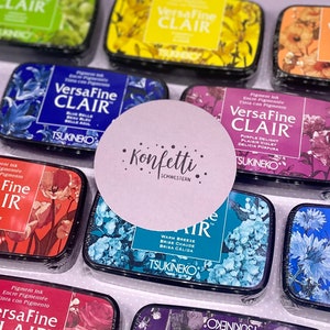 Könnte beinhalten: Eine Sammlung von bunten Stempelkissen mit floralen Designs und dem Text "VersaFine Clair" auf jedem Kissen. Die Stempelkissen sind in einem Raster auf einer weißen Oberfläche angeordnet. Der Text "Konfetti Schwestern" ist auf einem rosa runden Aufkleber sichtbar.