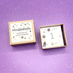 May include: A brown box with a white label that reads "Adventskalender 24 K&auml;rtchen mit positiven Gedanken". The box is open and a small white card with the number "1" and gold glitter is visible.