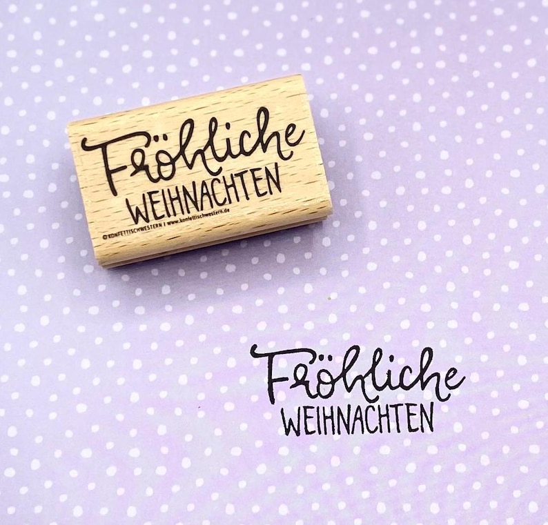 Peut inclure: Un tampon en bois avec le texte "Fr&ouml;hliche Weihnachten" en encre noire. Le tampon est sur un fond violet avec des points blancs.